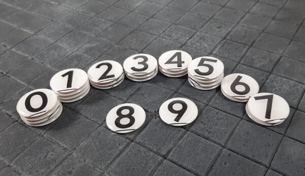 A collection of round, white tokens with black numbers ranging from 0 to 9 printed on them. The tokens are placed on a dark, tiled floor in stacks, with the numbers 0 through 7 neatly piled and the numbers 8 and 9 laying flat, all arranged in ascending order from left to right. The tokens feature a thin black line that underlines each number so that it is easy to tell which way is up.